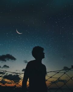 A lone silhouette stands beneath a crescent moon and starry sky, evoking a sense of solitude and wonder.