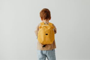 Cute child with red hair wearing a playful lion backpack facing away.